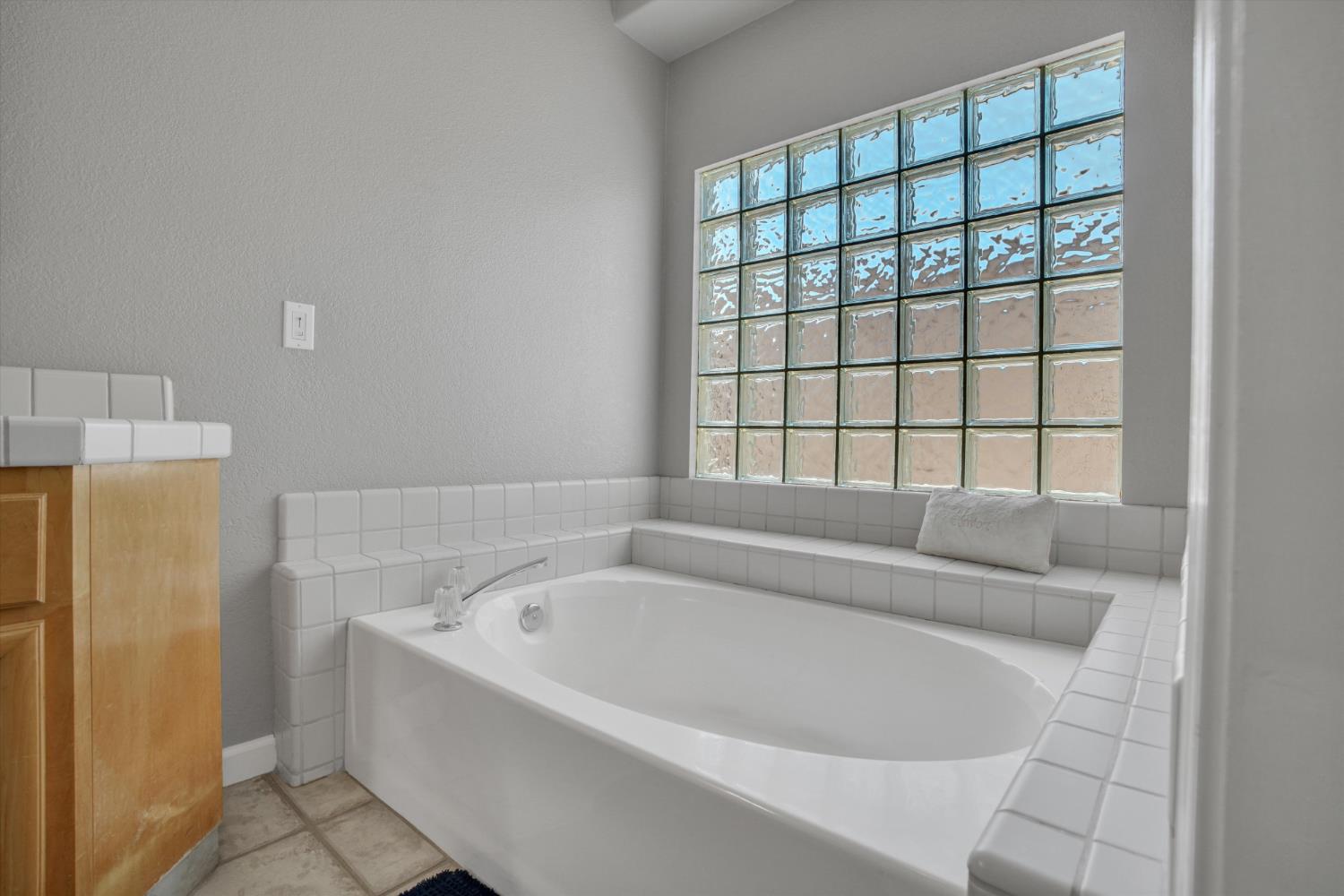 Bathroom with a large white soaking tub beneath a glass-block window and tiled surround.