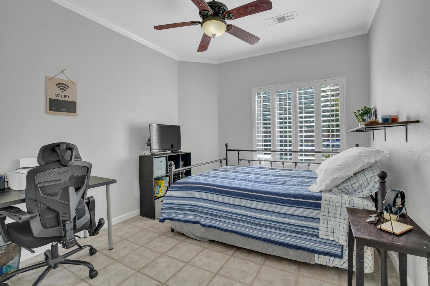 Bedroom with a blue-striped bed, plantation shutters, ceiling fan, and desk chair.