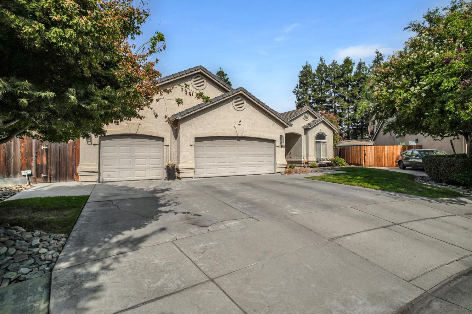 Beige stucco home with two garage doors, a wide driveway, and mature trees.