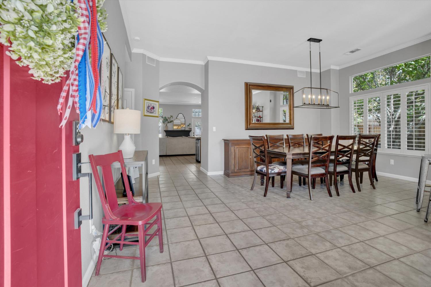 Dining room with a long wooden table, cross-back chairs, tiled floors, and plantation shutters.