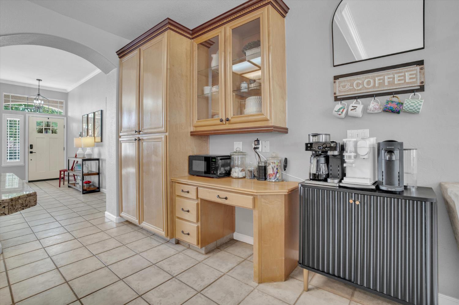 Kitchen coffee station with several coffee makers, light wood cabinets, and a tiled floor.
