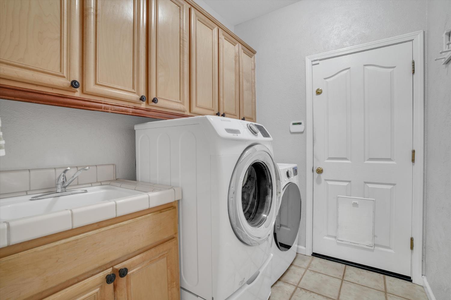 Laundry room with front-loading washer and dryer, utility sink, wood cabinets, and a pet door.