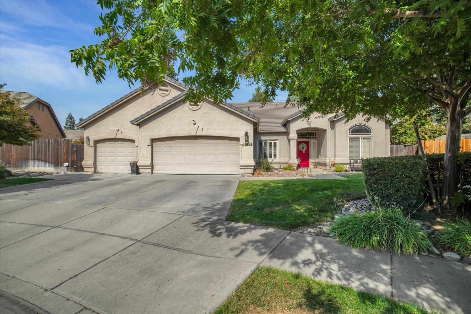Stucco home with two garage doors and a red front door beneath mature trees.