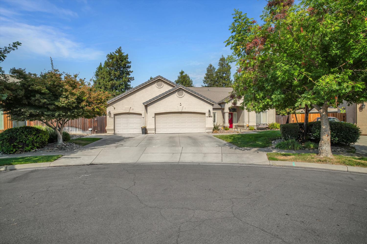Single-story stucco home with a two-car garage, red front door, and mature trees.