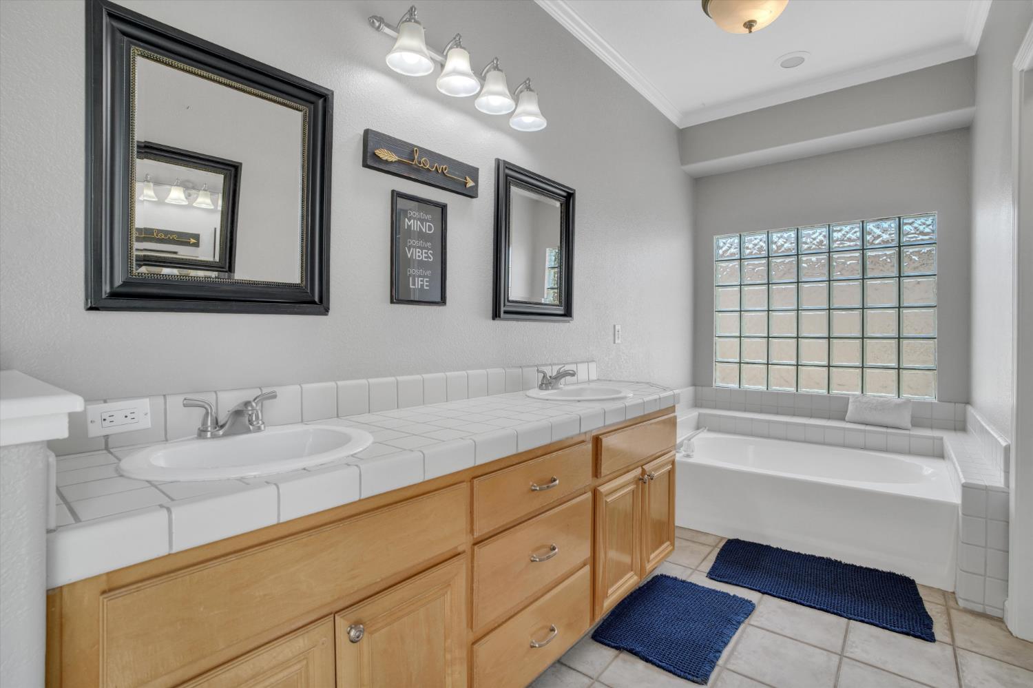 Bathroom with a double-sink tiled vanity, wood cabinets, and a bathtub beneath a glass-block window.