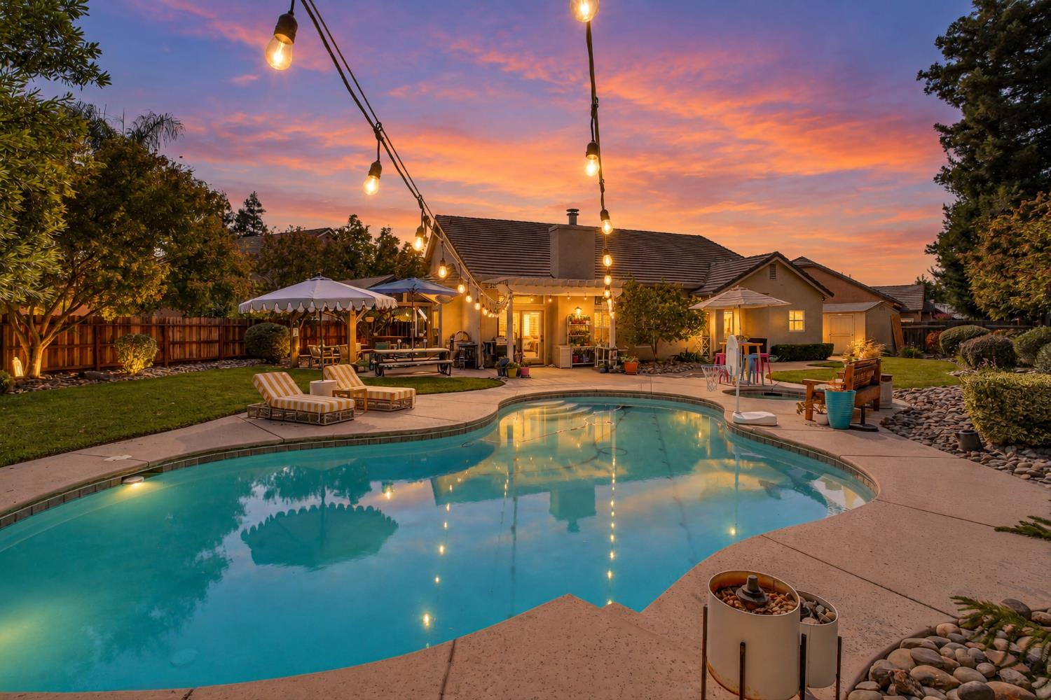 Backyard pool and patio at dusk with string lights and a colorful sunset behind the home.