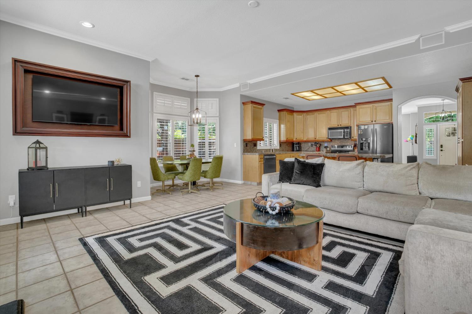 Open-plan living room and kitchen with a gray sectional, geometric rug, light wood cabinets, and stainless steel appliances.