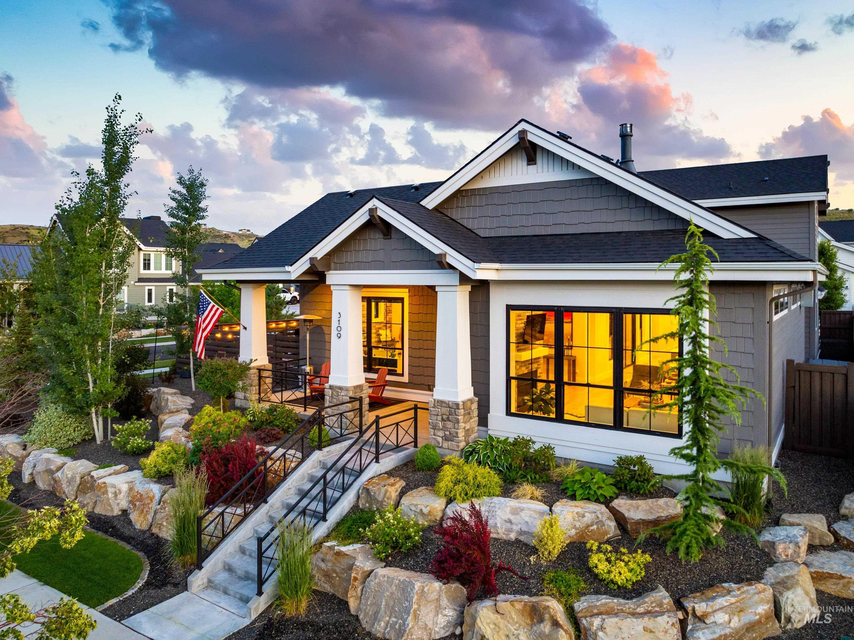 Gray shingle-sided home with a covered porch, warm-lit windows, and landscaped stone garden beds.