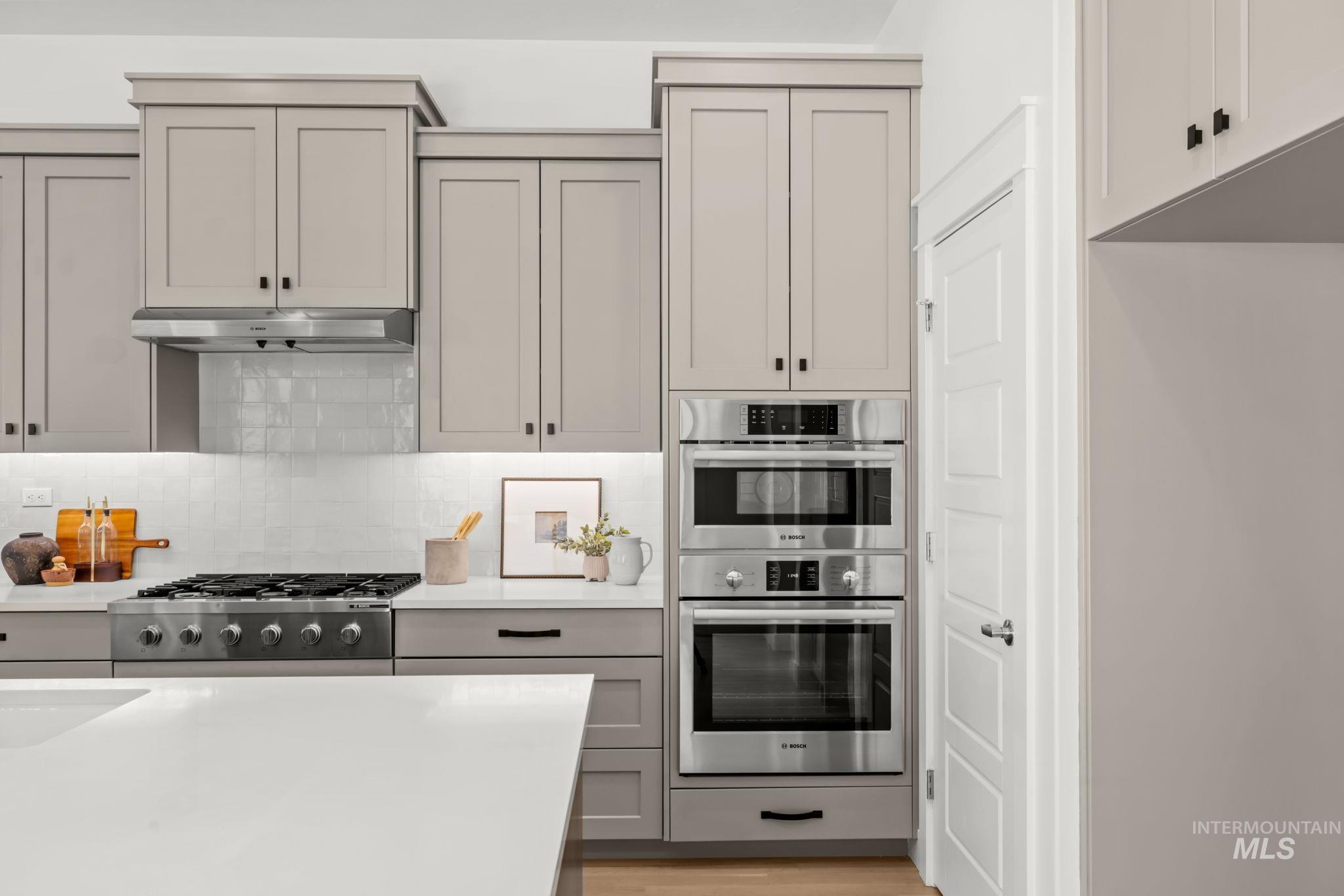 Kitchen with a white island, greige cabinets, gas range, and stacked stainless ovens.