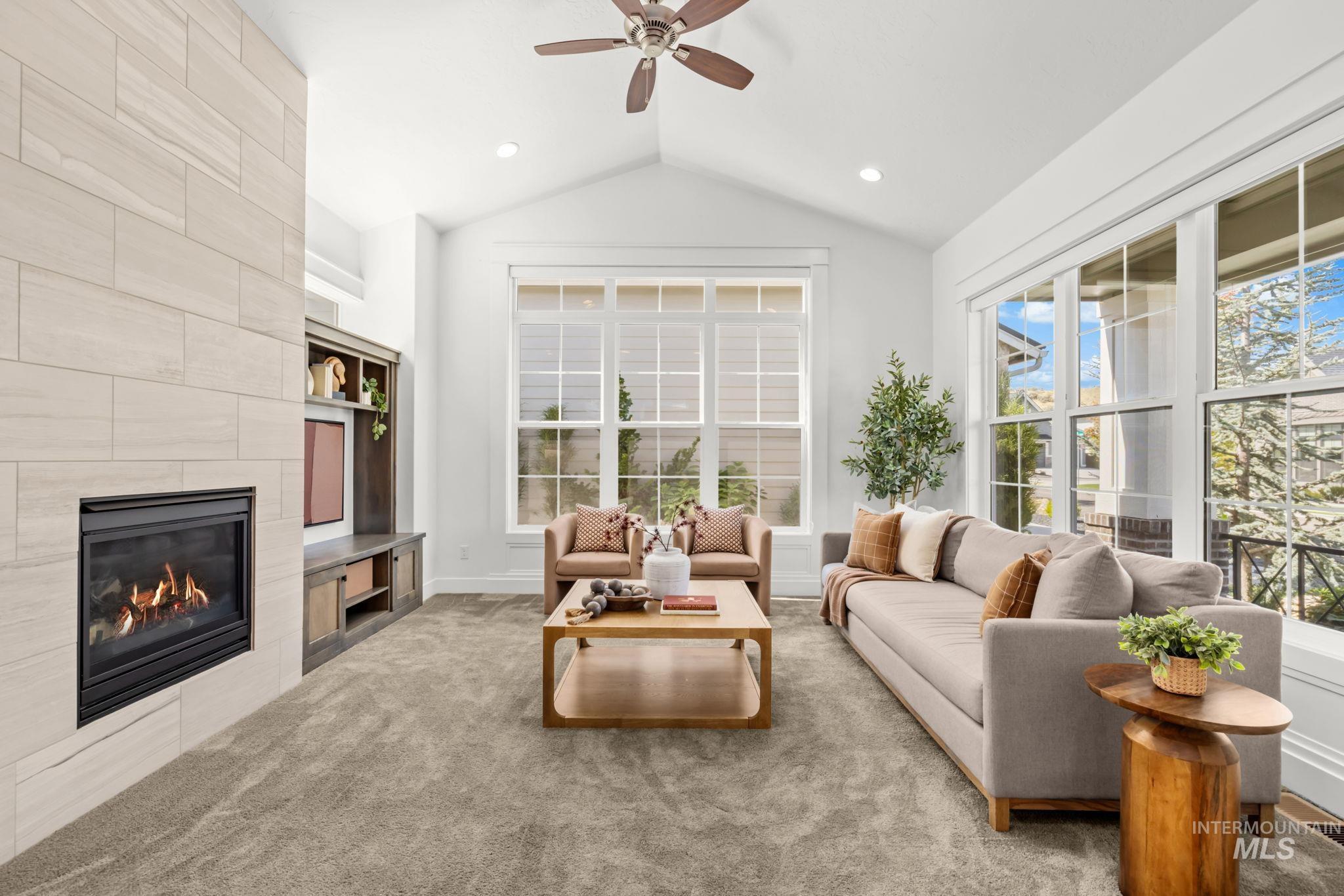 Bright living room with vaulted ceiling, tall windows, tiled fireplace, and neutral furnishings.