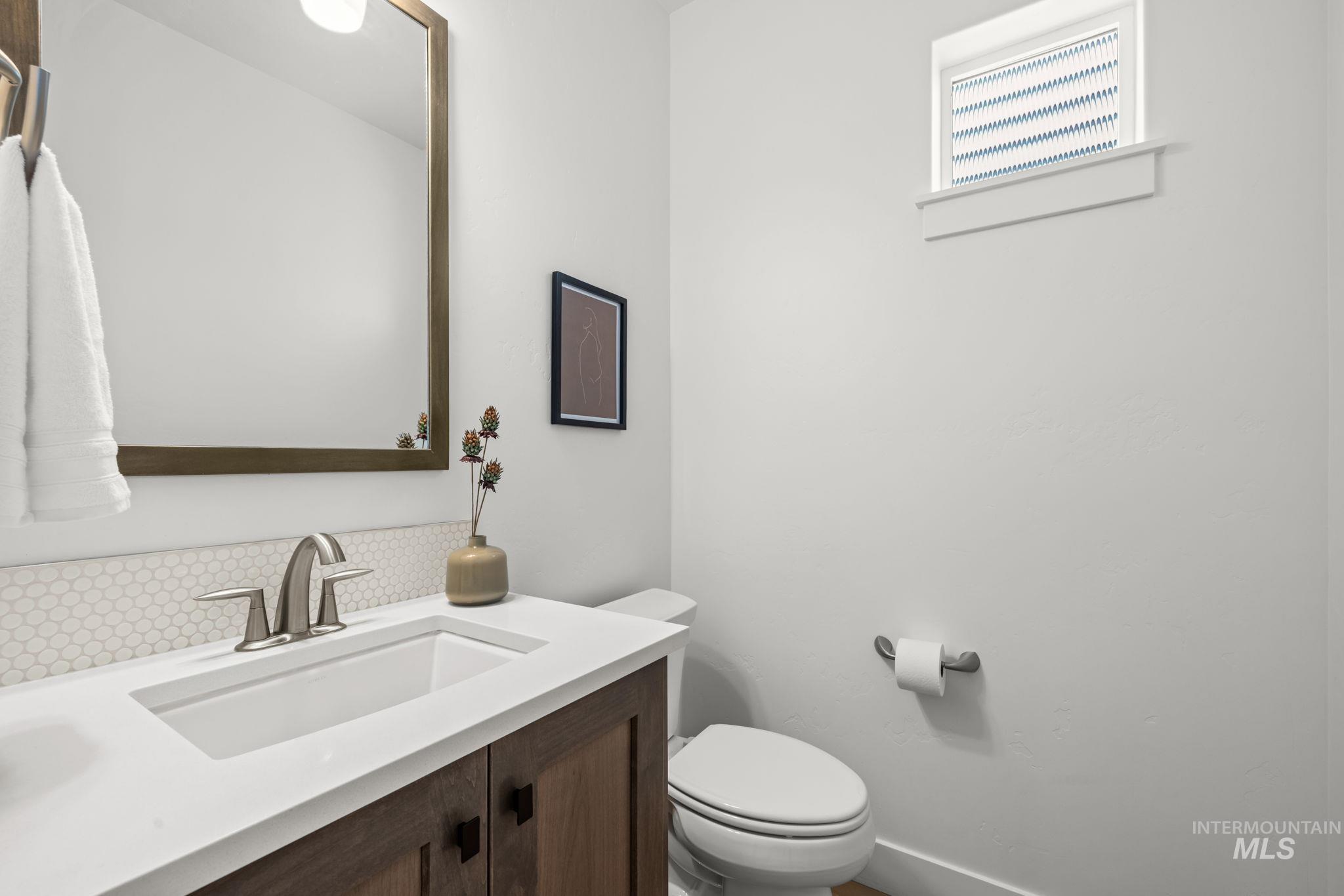 Bathroom with dark wood vanity, white sink, round-tile backsplash, and toilet beneath a high window.