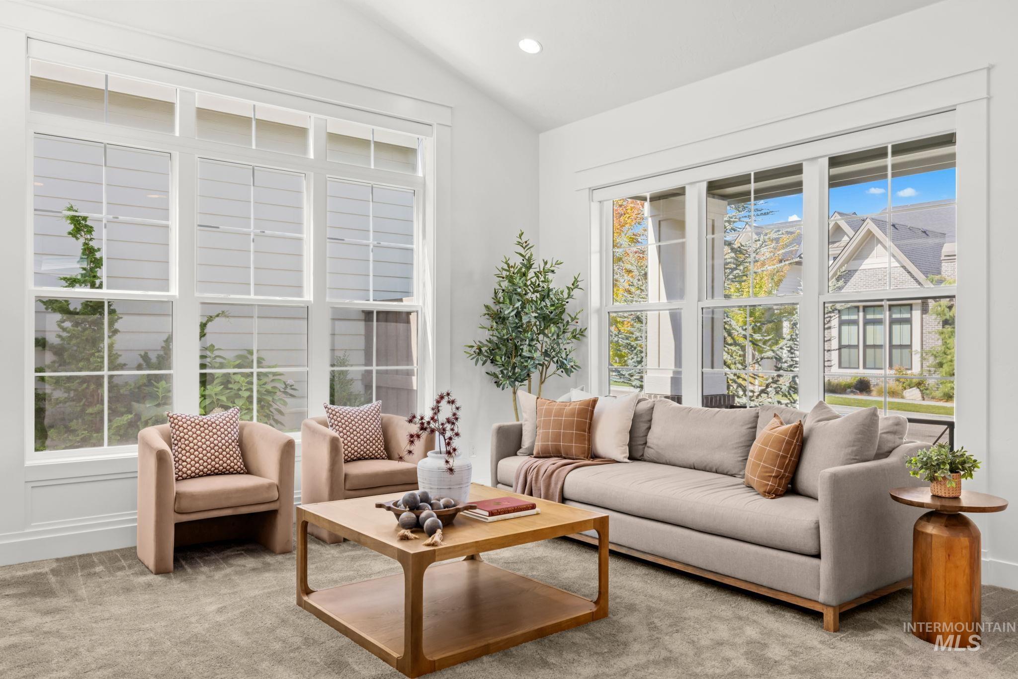 Furnished living room with a gray sofa, blush armchairs, and large windows overlooking autumn trees.