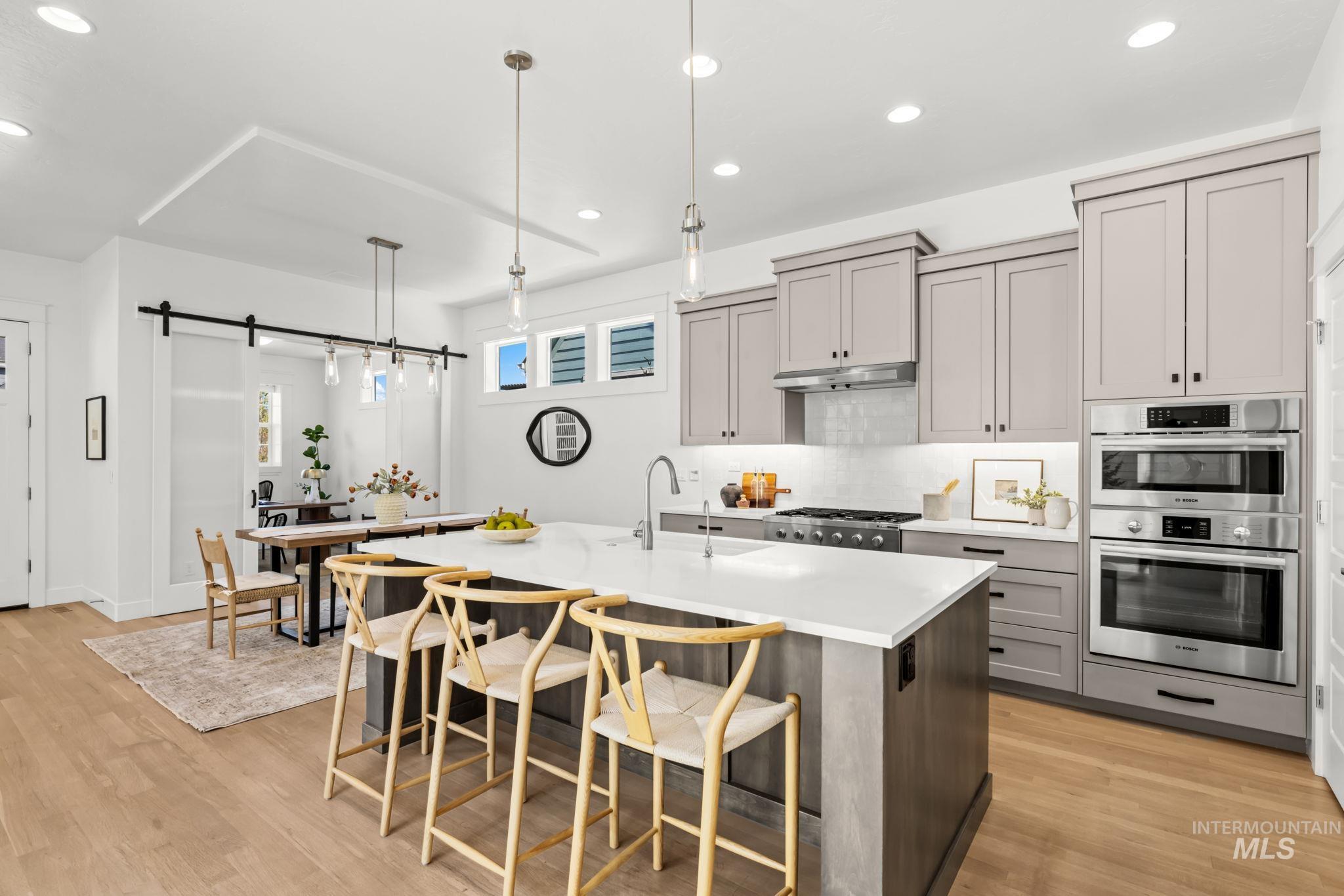 Kitchen with a large white island, light wood bar stools, gray cabinets, and double wall ovens.