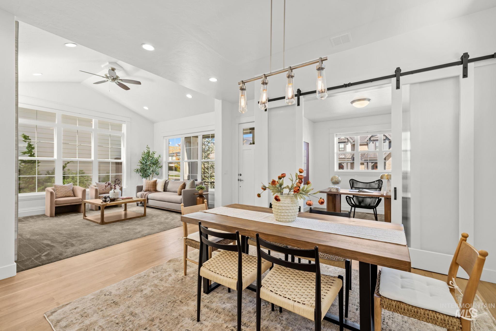 Open-plan dining and living area with a wood table, pendant lights, and a carpeted sitting area by large windows.