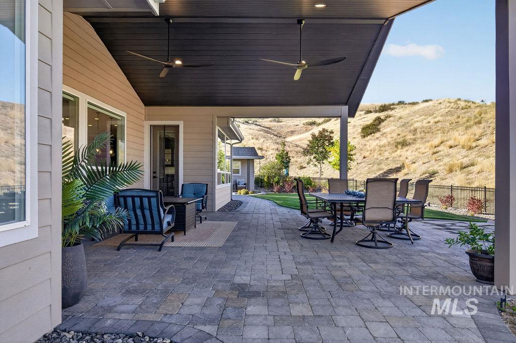 Covered patio with stone pavers, ceiling fans, lounge seating, and a dining table overlooking grassy hills.