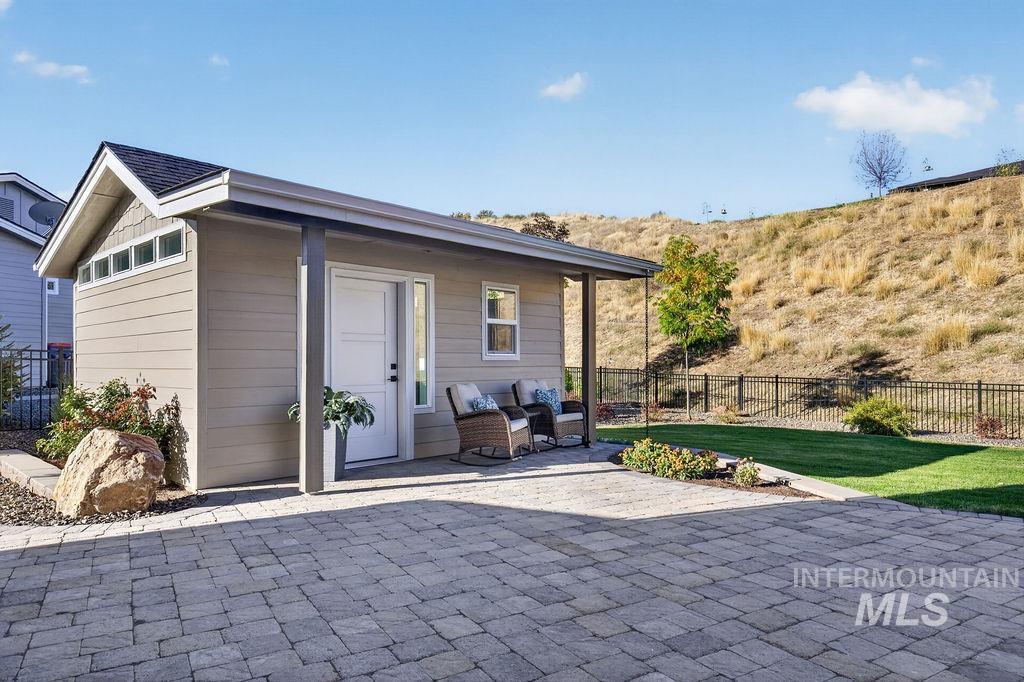 Covered patio with two chairs beside a gray-sided structure, overlooking a lawn and dry hillside.