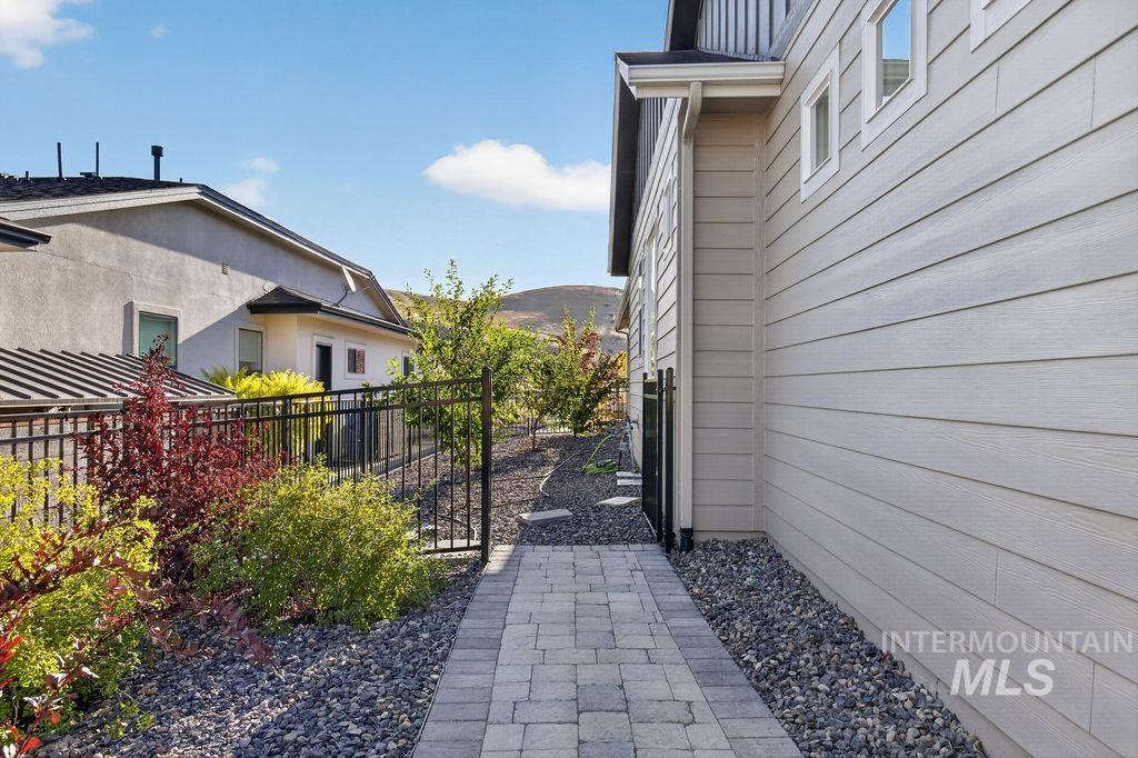 House side yard with a paved walkway, black metal fence, gravel landscaping, and distant hills.