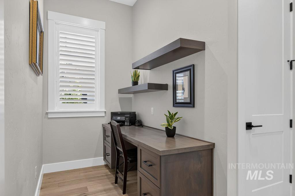Home office with a gray wood desk, floating shelves, and a window with plantation shutters.