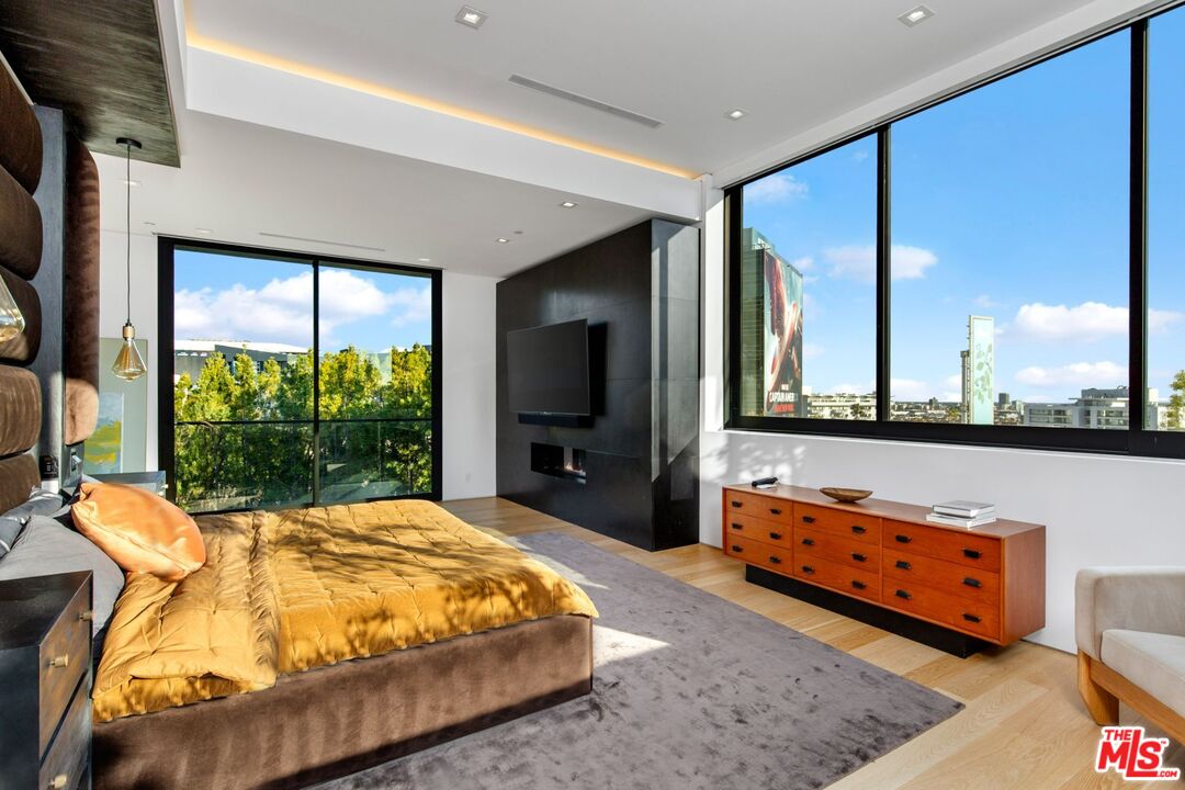 Modern bedroom with a mustard-gold quilted bed and large windows overlooking trees and city buildings.