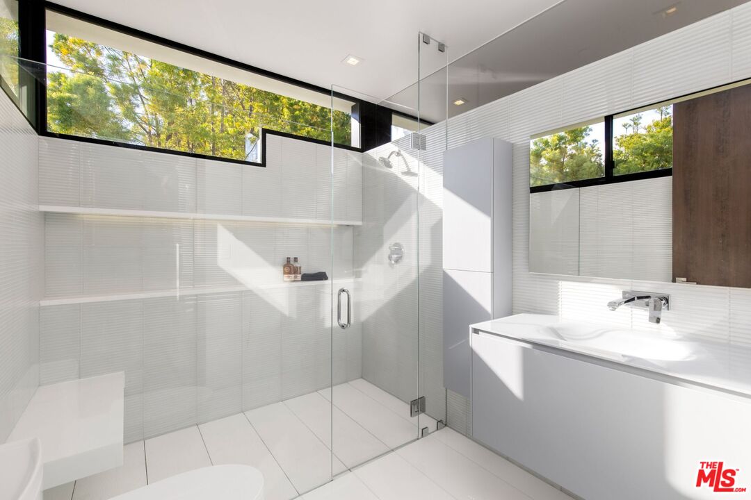 Modern white bathroom with a glass shower, high windows, and a wide mirrored vanity.