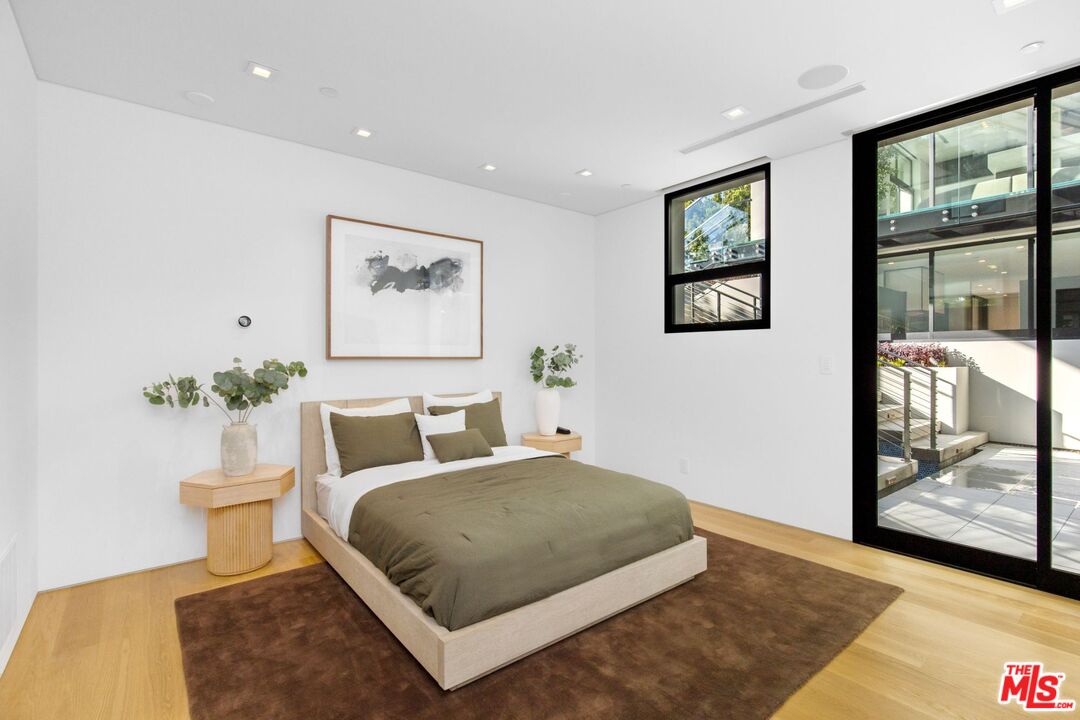 Modern bedroom with olive bedding, light wood furnishings, and black-framed glass doors to a patio.