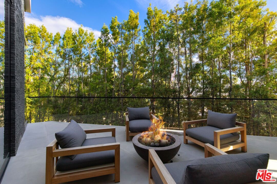 Patio lounge chairs surround a fire pit, with a wooded view beyond the glass railing.