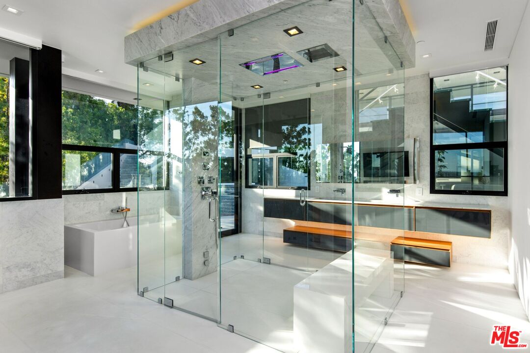 Modern bathroom with a glass-enclosed shower, marble surfaces, soaking tub, and wood-accented vanity benches.
