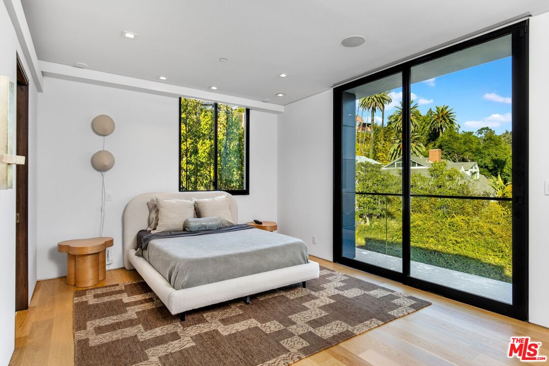 Modern bedroom with a low upholstered bed and large glass doors overlooking leafy trees.