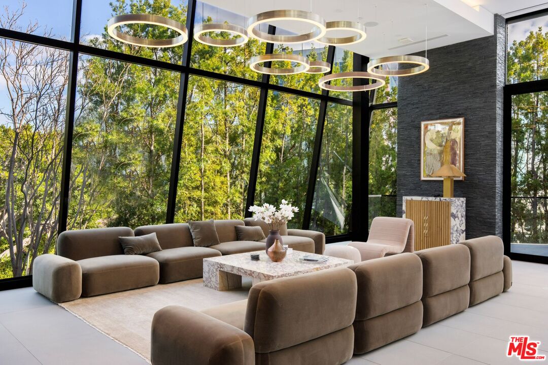 Contemporary living room with rounded taupe sofas, circular pendant lights, and floor-to-ceiling windows overlooking trees.
