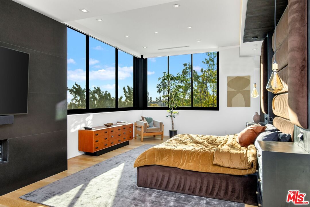 Modern bedroom with gold bedding and floor-to-ceiling corner windows overlooking trees.