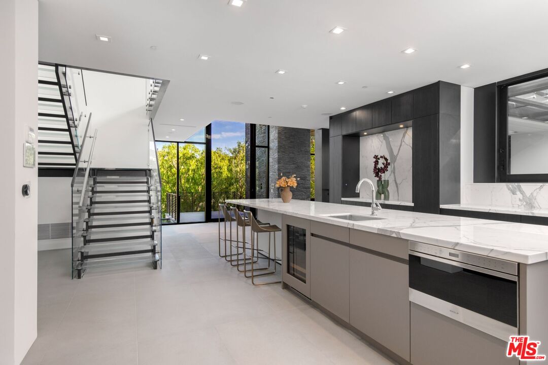Modern kitchen with a marble island, dark cabinetry, and an open staircase beside floor-to-ceiling windows.