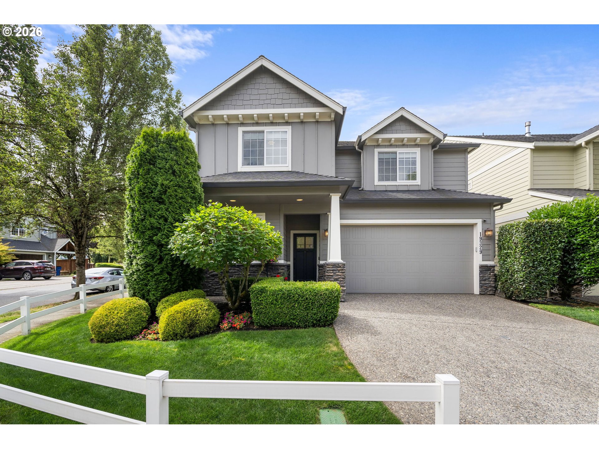 Gray two-story house with a two-car garage, landscaped front yard, and white fence.