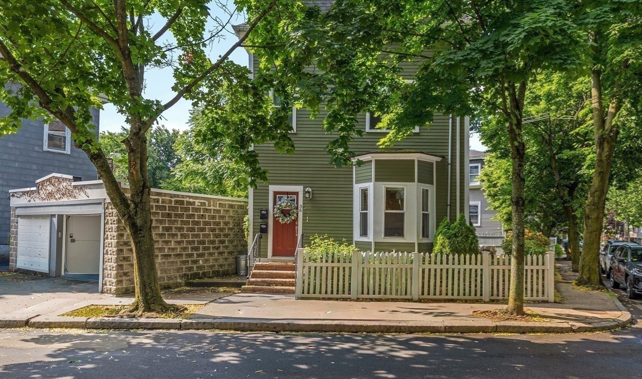 Green-sided Somerville home with a bay window, red front door, and white picket fence beneath leafy trees.