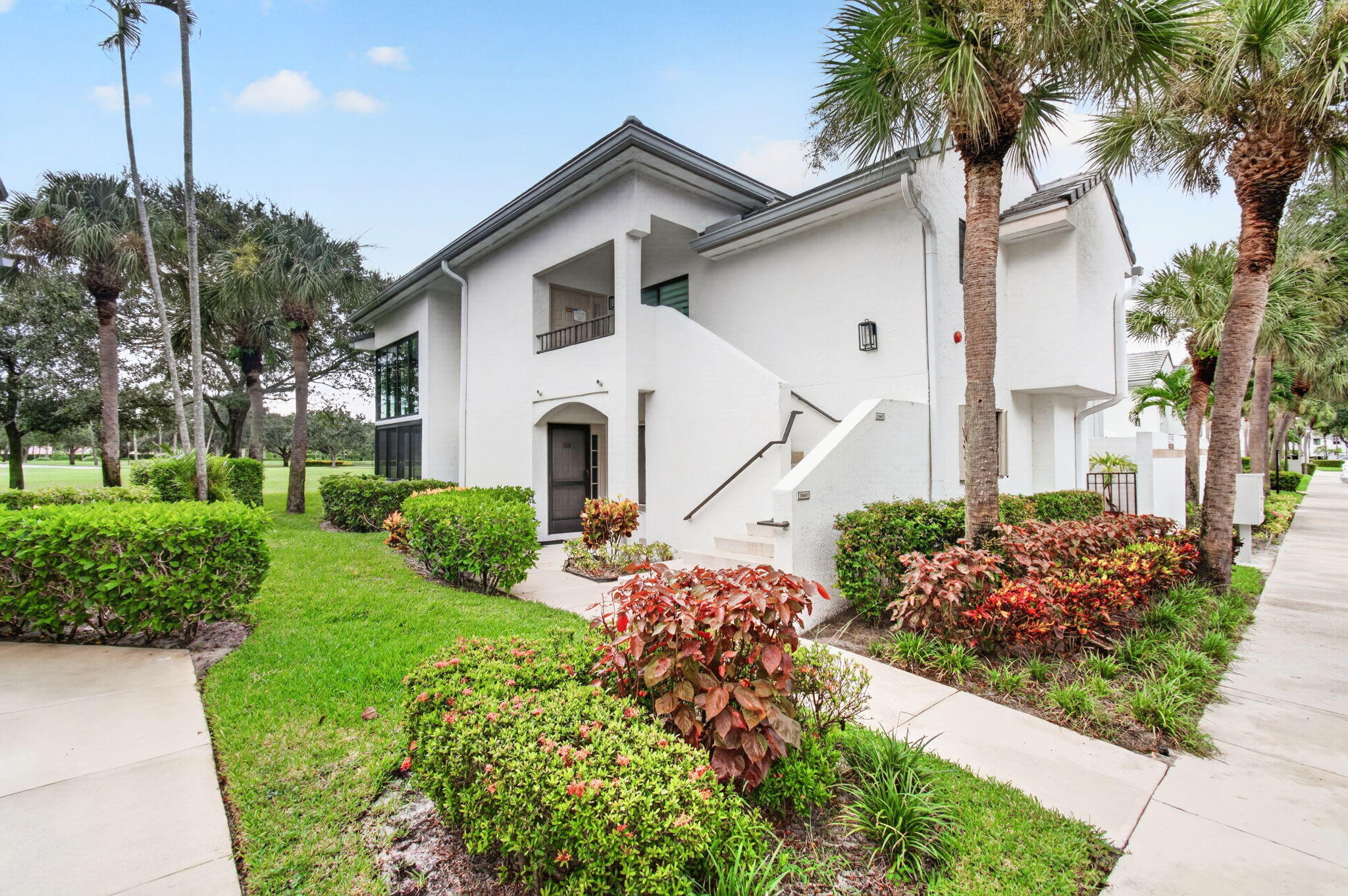 White two-story home with an exterior staircase, framed by palm trees and landscaped gardens.