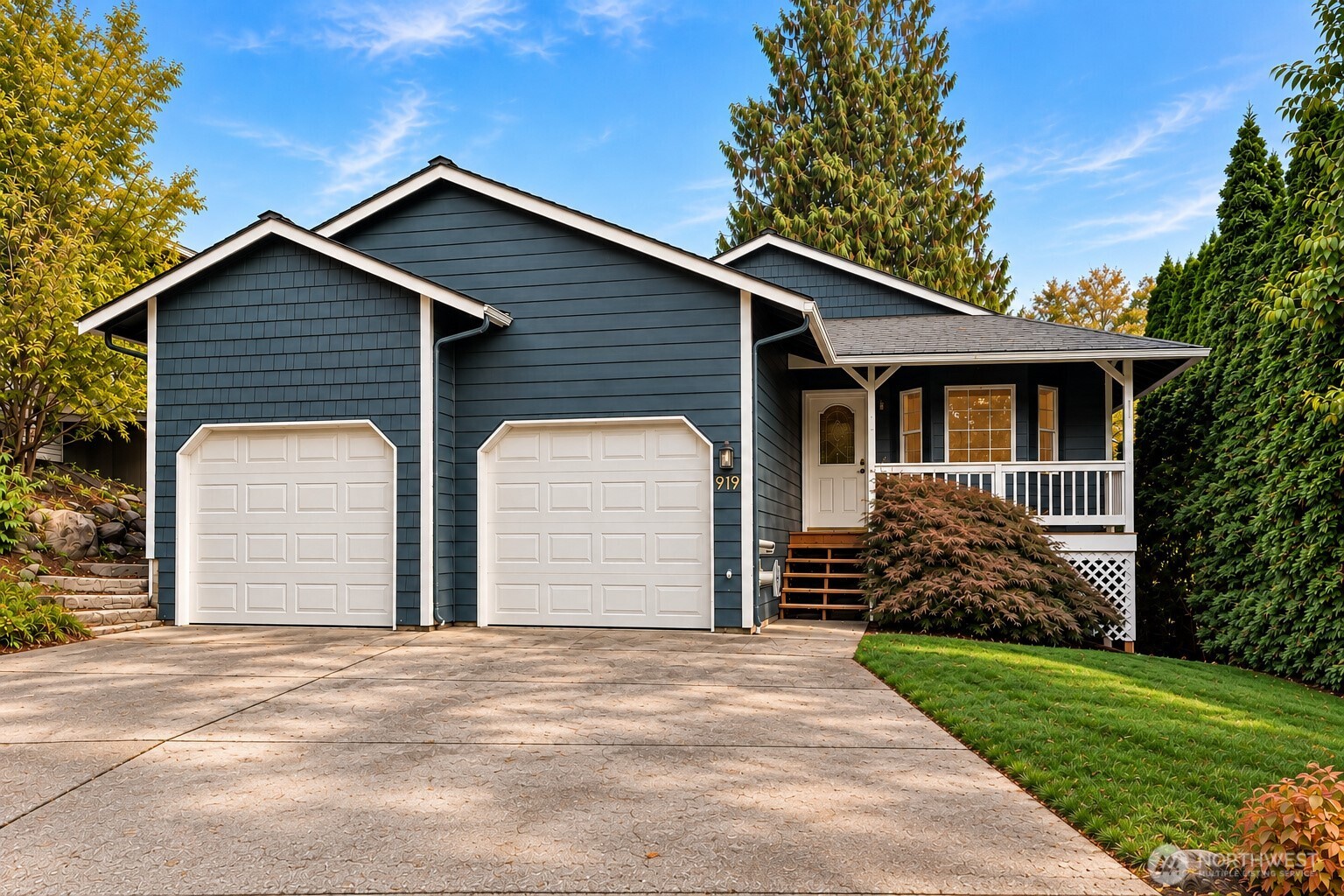 Dark blue house with two white garage doors, a covered porch, and a broad paved driveway.
