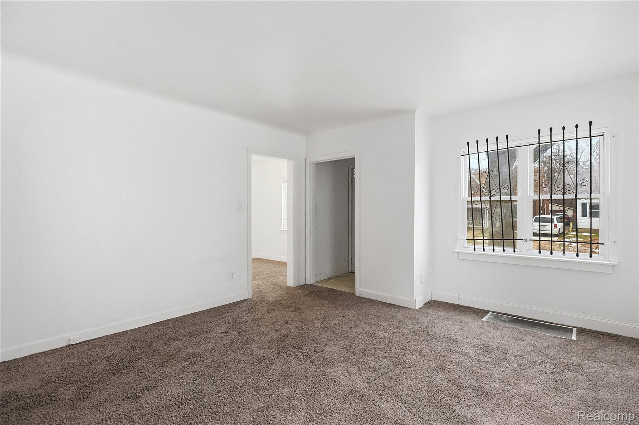 Empty carpeted room with white walls and a window fitted with decorative security bars.