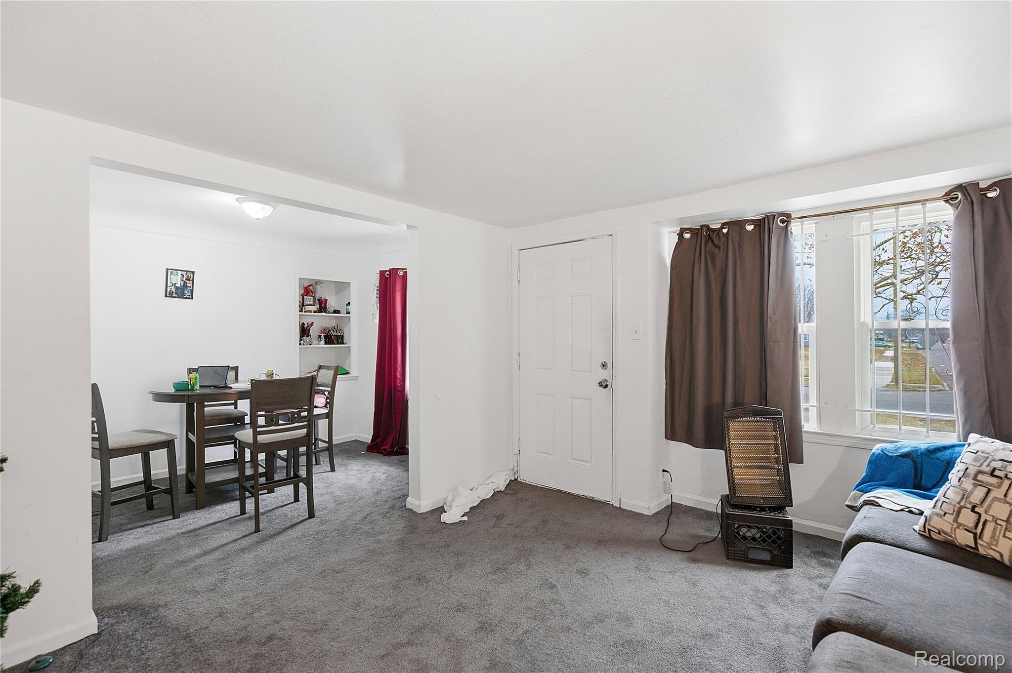 Living room with gray carpet, a gray sofa, and a large curtained window beside an adjoining dining area.