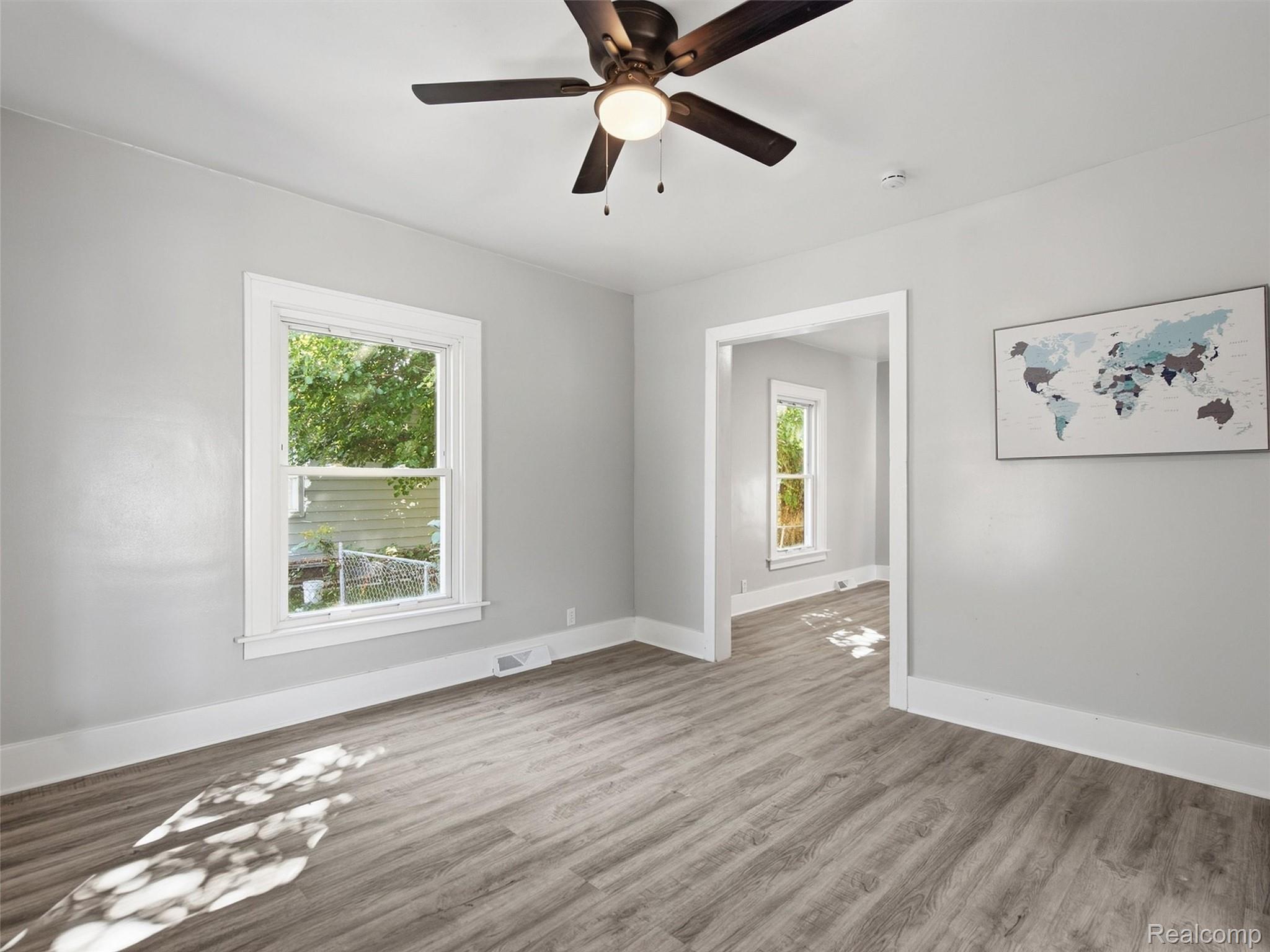 Empty gray room with light wood-look floors, ceiling fan, framed world map, and doorway to an adjoining room.
