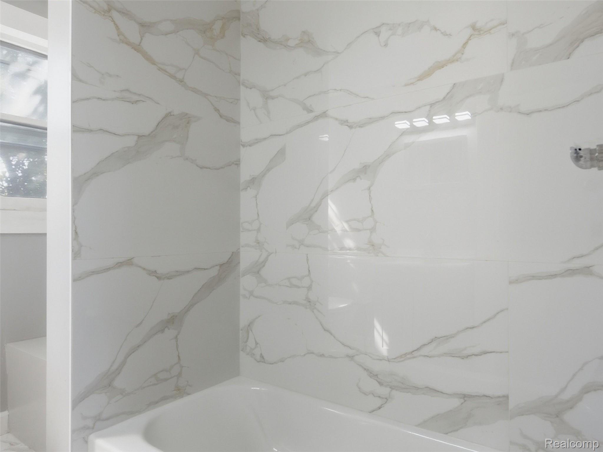 Bathroom with a white bathtub and marble-look tile surround beside a window.