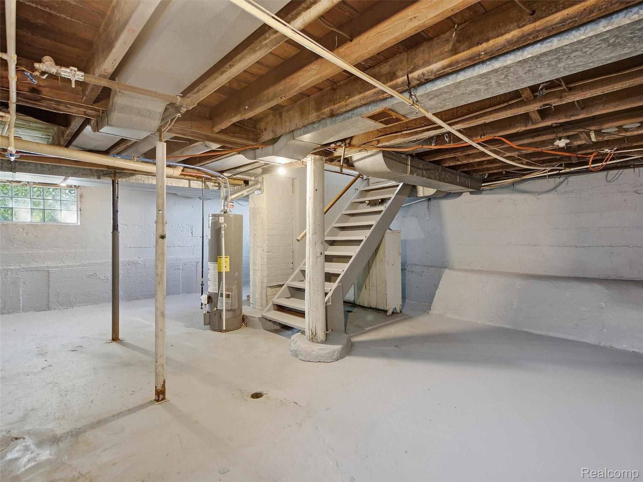 Unfinished basement with exposed ceiling joists, a staircase, water heater, and glass-block window.