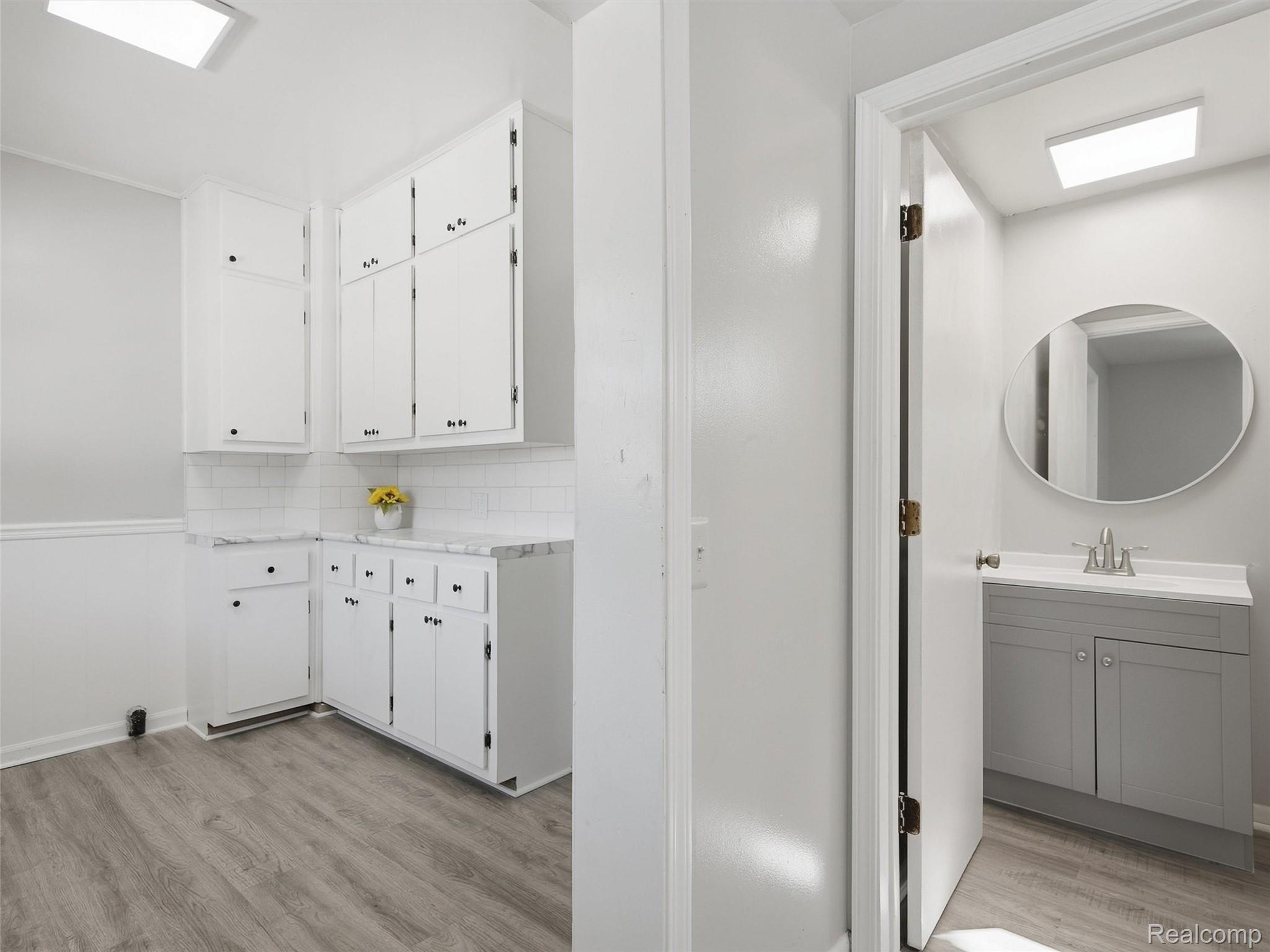 Laundry room with white cabinets and gray flooring, opening to a bathroom with a gray vanity and oval mirror.