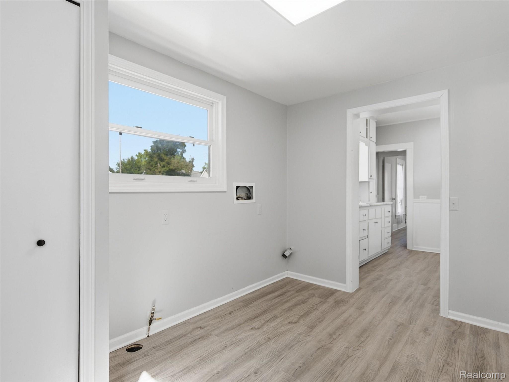 Empty room with wood-look flooring, a large window, and exposed plumbing beside a doorway to a white vanity.
