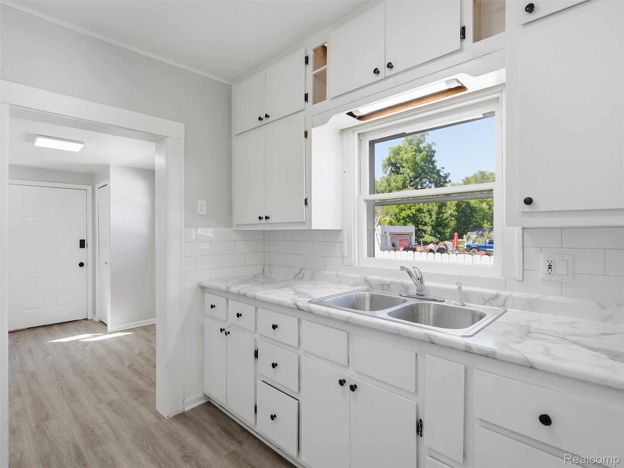 Bright white kitchen with marble-look counters, a double sink, and a window overlooking trees.