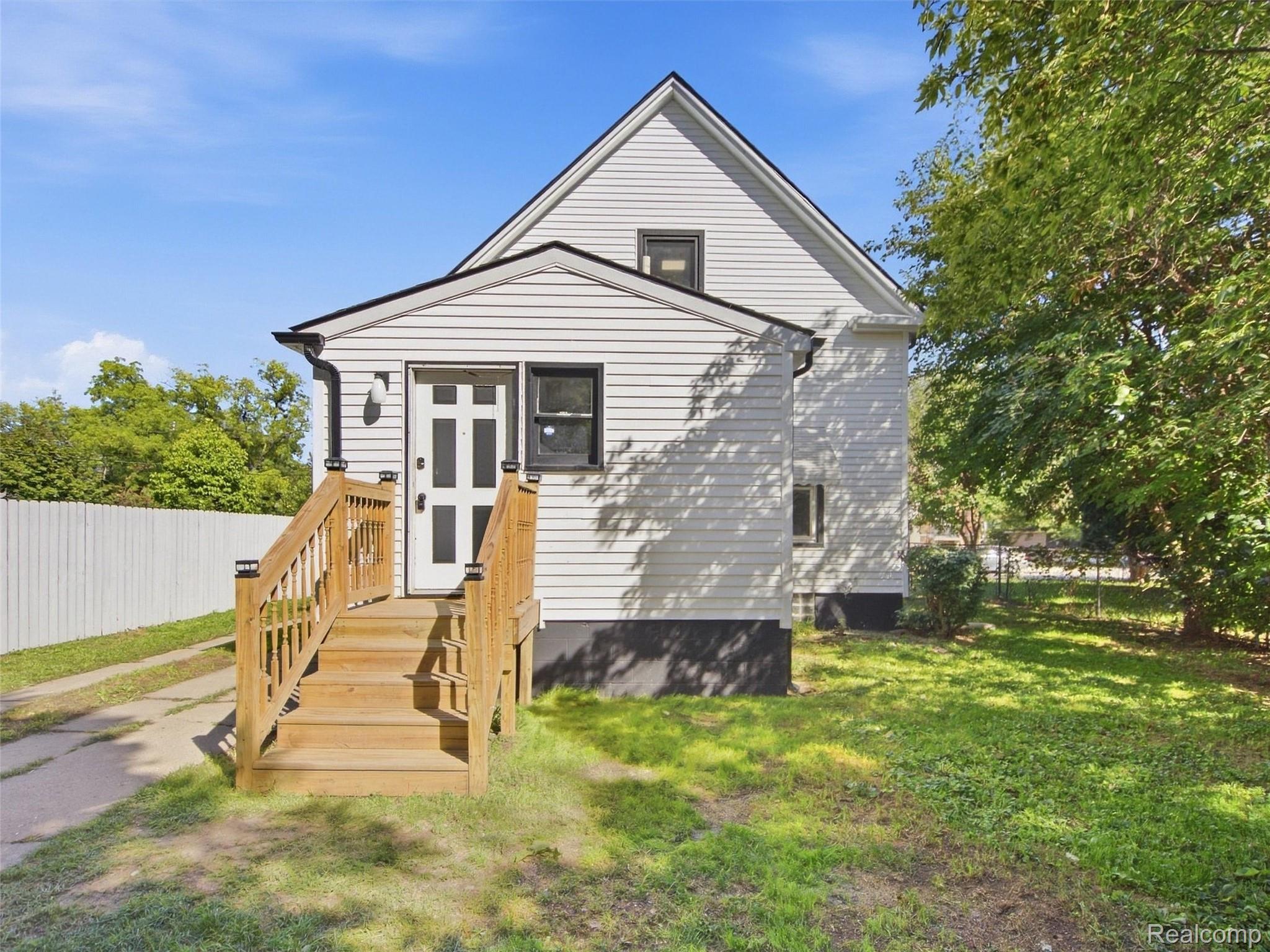 White gable-roofed house with a raised wooden entrance, white fence, and leafy yard.