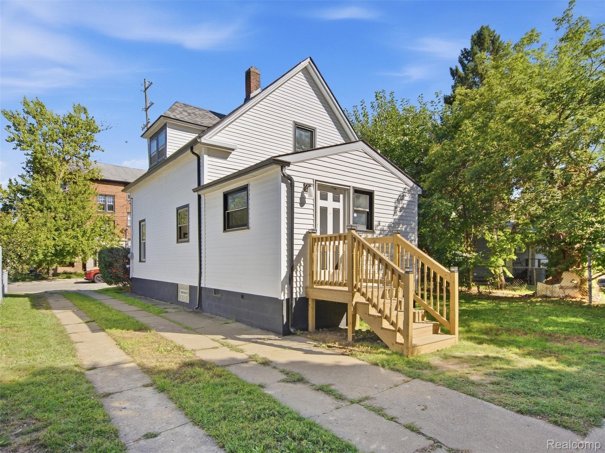 White two-story house with a steep gabled roof and wooden front stairs beside a concrete sidewalk.