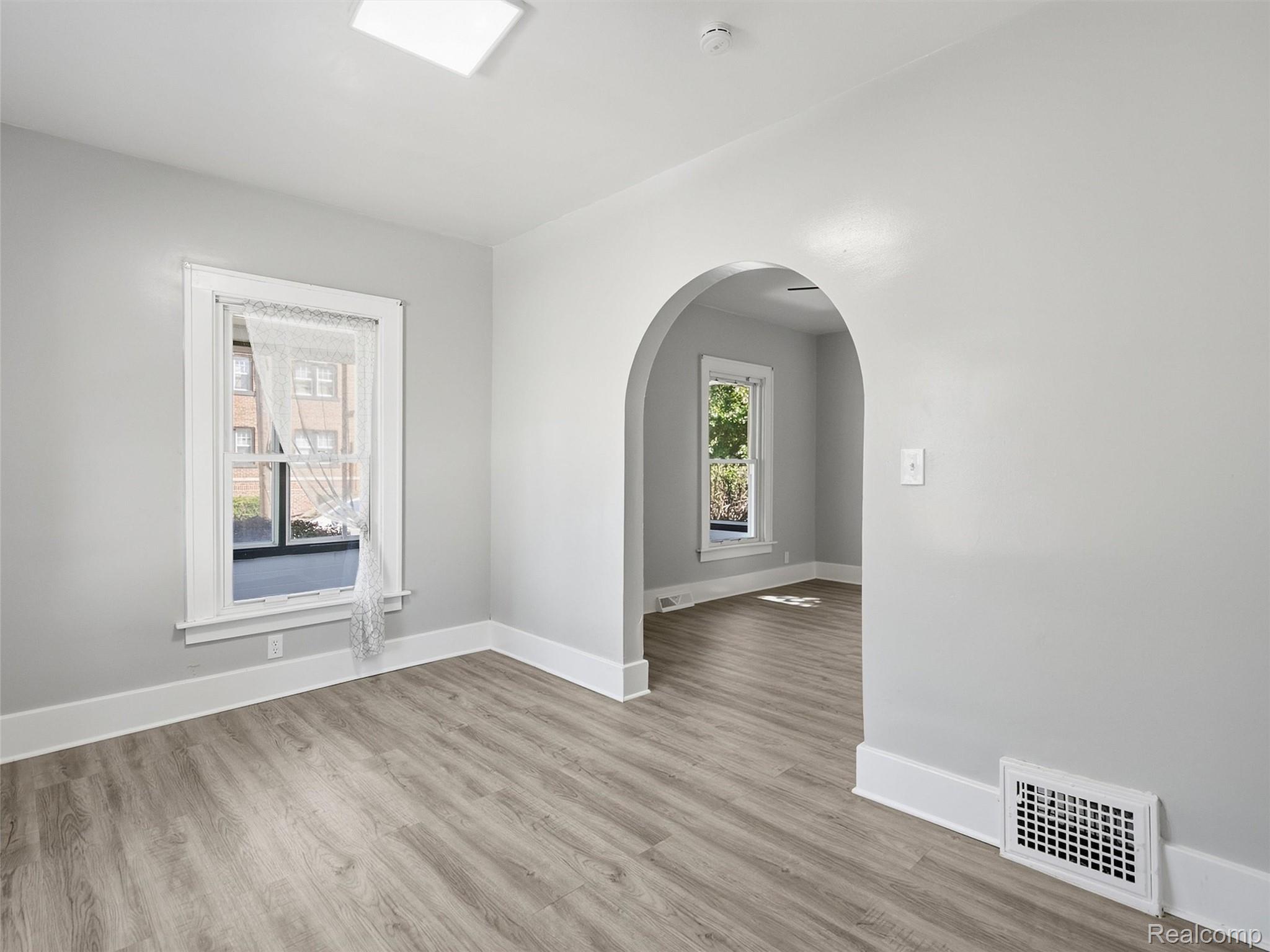 Vacant room with gray wood-look flooring, a curtained window, and an arched opening to a second room.