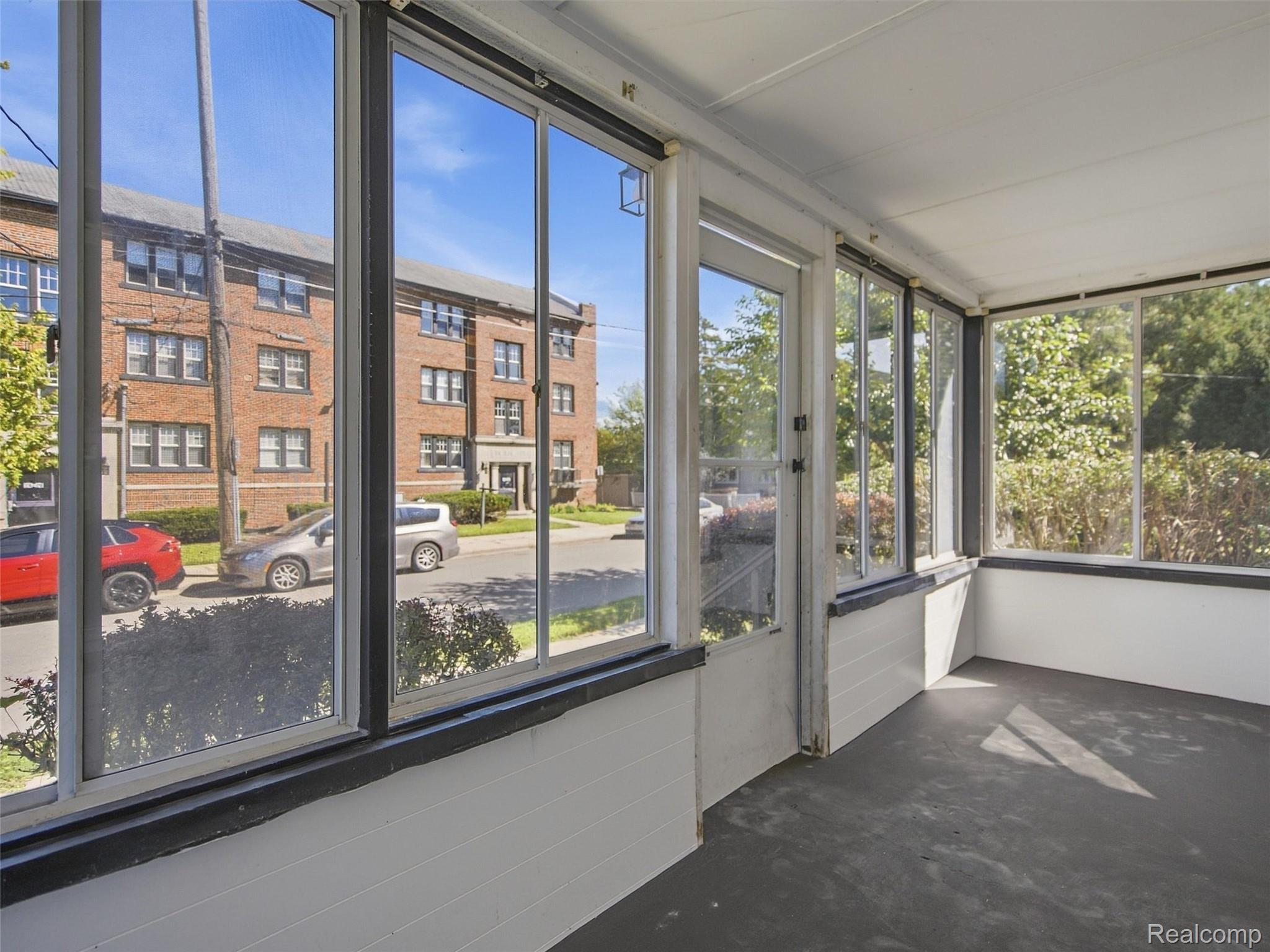 Empty enclosed porch with large windows overlooking a leafy street and red-brick apartment building.