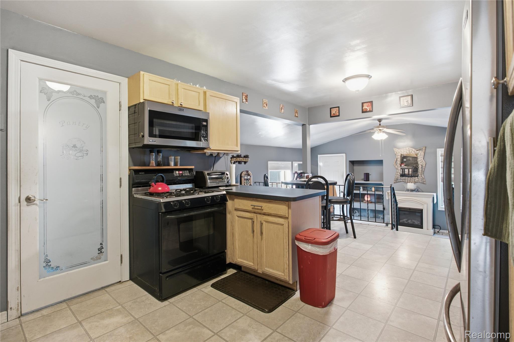 Kitchen with light wood cabinets and black appliances, opening into a dining area with a fireplace.
