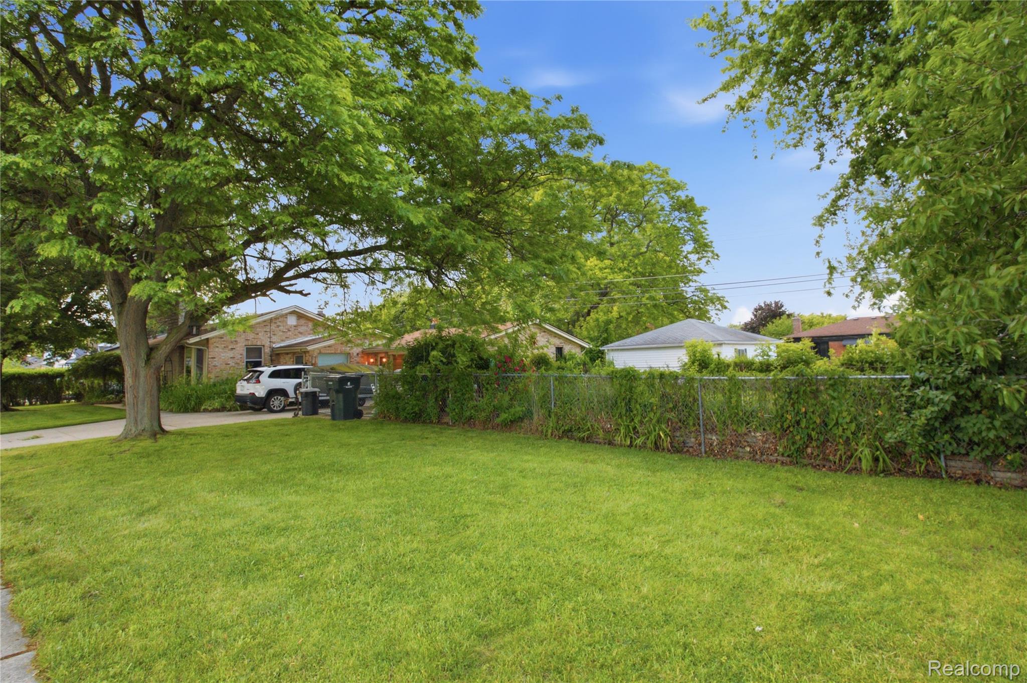 Brick house partly screened by mature trees beyond a fenced lawn and driveway.