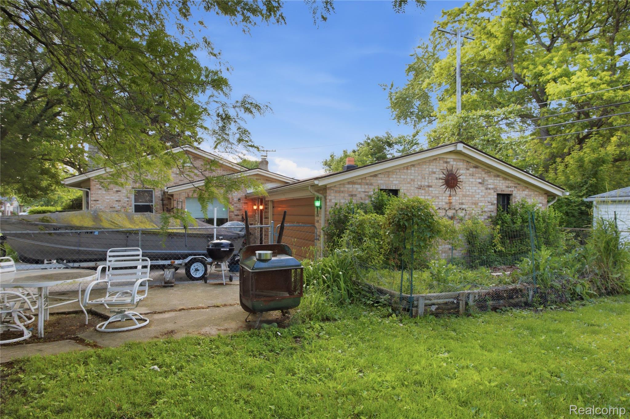 Brick home’s rear exterior with a patio, boat on a trailer, and fenced garden beds.